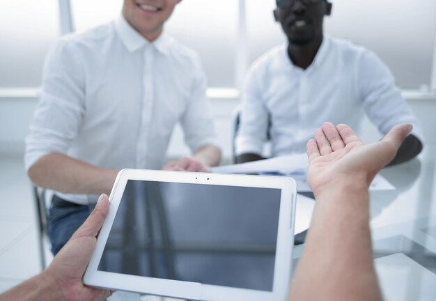 A close-up of an employee's hands selecting different voluntary benefits options on a tablet device. The background shows a blurred image of colleagues collaborating in an office setting.