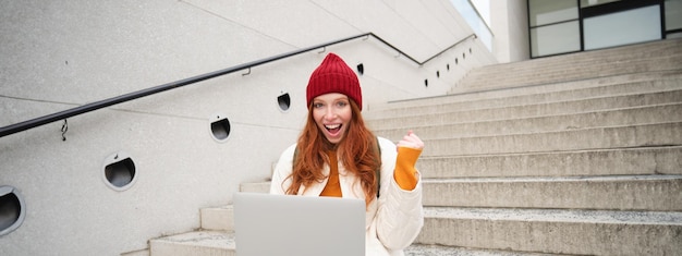 A student excitedly checking their email on a laptop, with a notification showing they received a scholarship. The background is a celebratory collage of graduation photos.